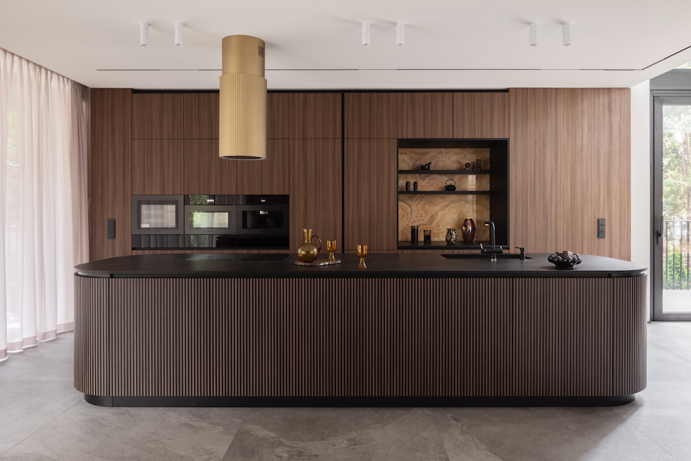 Elegancka kuchnia z dużą, ryflowaną wyspą, czarnym blatem i wysoką zabudową z forniru drewnianego. Nad wyspą wisi złoty, cylindryczny okap. W zabudowie widoczny piekarnik i ekspres do kawy.