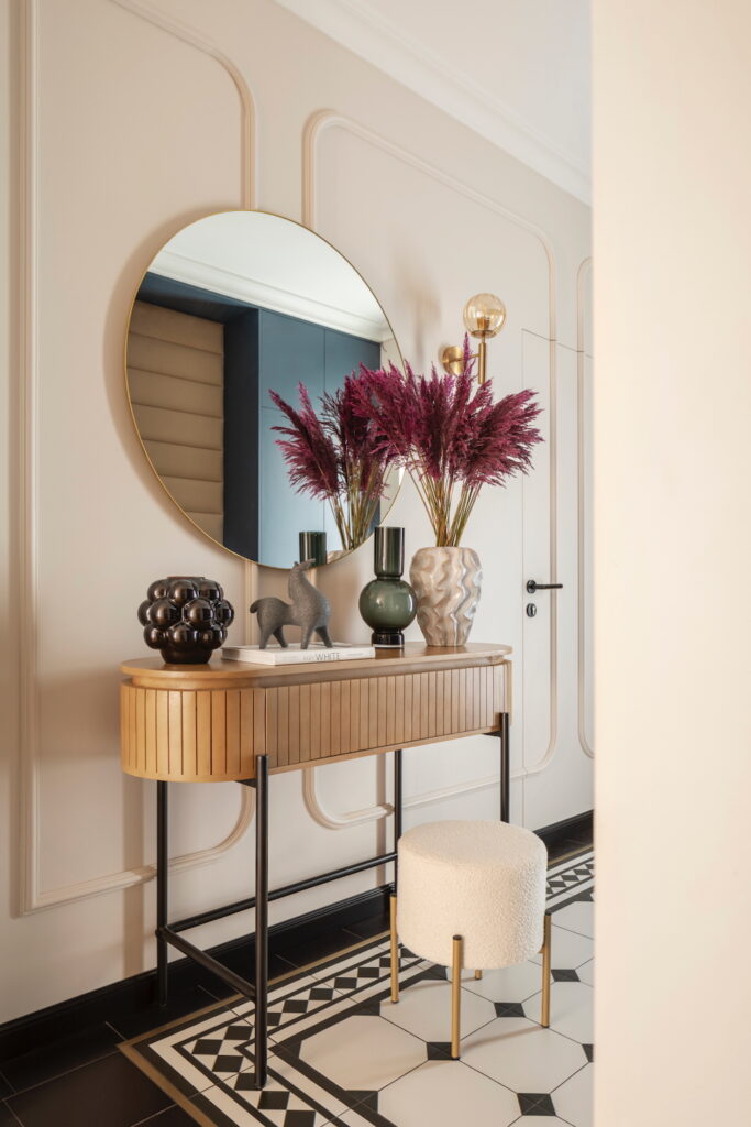 Stylowa konsola z lamelami, okrągłe lustro i bordowe trawy dekoracyjne w holu urządzonym przez homestagera.